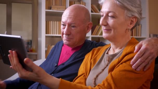 Profilbild av Senior kaukasiska makar titta på tabletten är roat sitter vid soffan i vardagsrummet. — Stockvideo