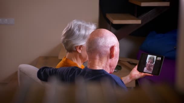 Plan arrière du couple caucasien aux cheveux gris senior ayant un vidéocall en ligne avec leur ami au téléphone à la maison confortable . — Video