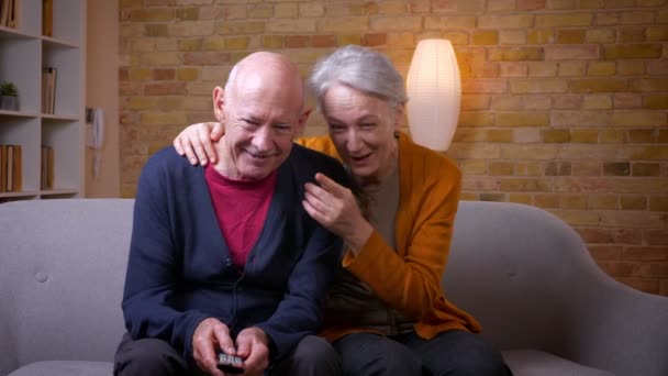 Senioren kaukasischen Ehegatten sehen Komödie im Fernsehen streicheln einander und lachen über Sofa im Wohnzimmer. — Stockvideo