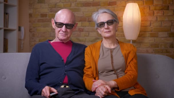 Senior caucásico cónyuges en 3d gafas ver película en la televisión abrazándose unos a otros en sofá en sala de estar . — Vídeos de Stock