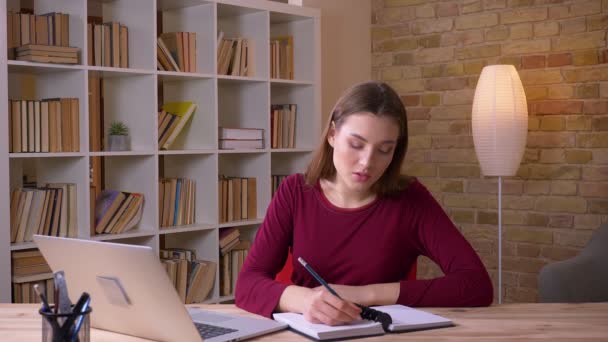 Junge brünette langhaarige Geschäftsfrau, die mit Laptop arbeitet, macht sich im Büro konzentriert Notizen. — Stockvideo