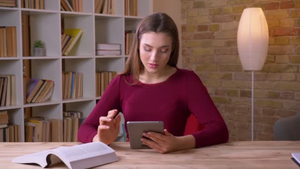 Jovem e bela morena estudante do sexo feminino trabalhando com tablet e livro sorri para a câmera no escritório . — Vídeo de Stock