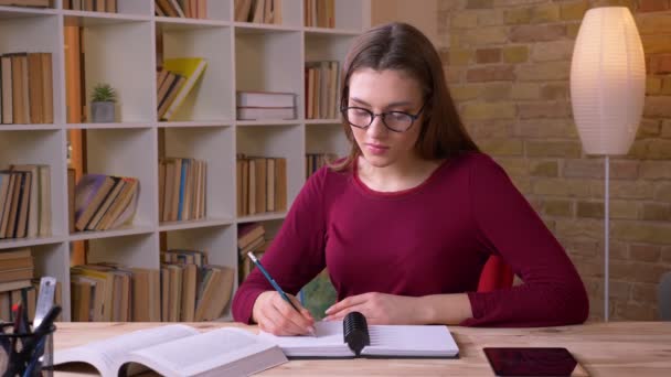 Jonge brunette langharige vrouwelijke student in bril lezen boek maakt notities oplettend in het kantoor. — Stockvideo