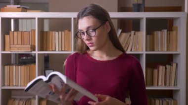 Genç ve güzel esmer, gözlüklü kız öğrenci kütüphanede kitap okurken kameraya gülümsüyor..