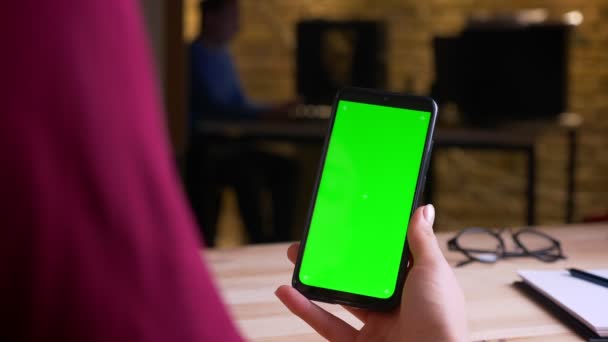 Tiro traseiro de mãos femininas segurando verticalmente smartphone com tela de croma verde no escritório . — Vídeo de Stock