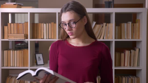 Joven y hermosa estudiante morena en gafas de ojo leyendo el libro con atención en la biblioteca . — Vídeo de stock