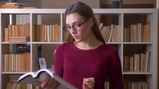 Young and beautiful brunette female student in eye glasses reading book watches into camera in library. — Stock Video