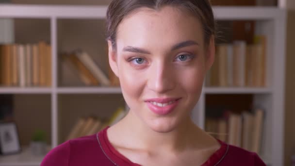 Portrait of young brunette female student with ponytail smiles modestly into camera in library. — Stock Video