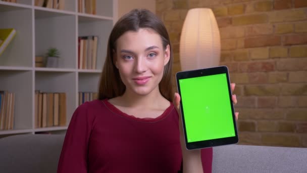 Junge und schöne brünette Frau zeigt aufrechten grünen Bildschirm des Tablets in die Kamera und empfiehlt die App zu Hause. — Stockvideo