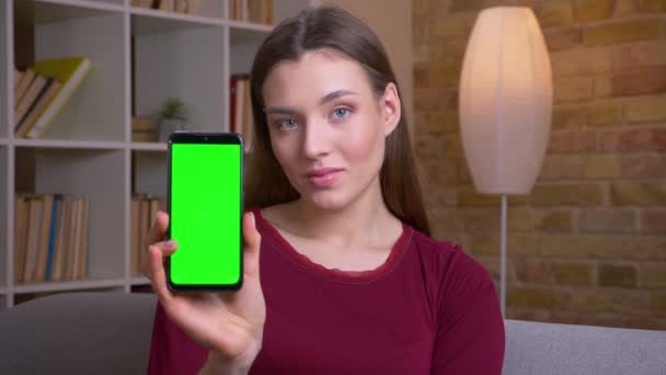 Young and pretty brunette female shows upright green chroma screen of phone recommending the app at home. — ストック動画