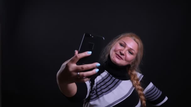 Retrato de primer plano de una mujer caucásica de mediana edad tomando selfies en el teléfono con fondo aislado en negro — Vídeo de stock