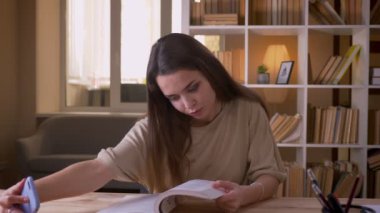 Genç çekici beyaz kız öğrencinin kitap okuyup ofiste kapalı mekanlarda telefonda selfie çektiği yakın çekim portresi
