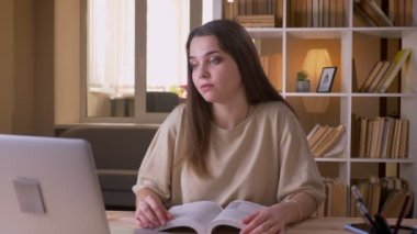 Genç çekici beyaz kız öğrencinin kitap okuması ve ofisteki dizüstü bilgisayarda görüntülü arama yaptığı closeup portresi