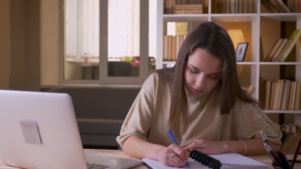 Primer plano retrato de joven atractiva mujer de negocios caucásica utilizando el ordenador portátil y tomando notas en el interior de la oficina — Vídeos de Stock