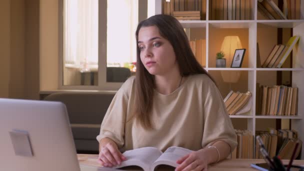 Retrato de cerca de una joven atractiva estudiante caucásica leyendo un libro y haciendo una videollamada en el portátil dentro de la oficina — Vídeos de Stock