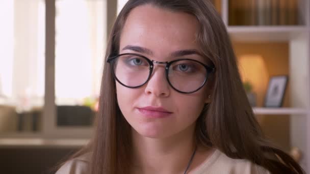 Крупним планом портрет молодої досить кавказької студентки в окулярах, дивлячись на камеру в приміщенні в квартирі — стокове відео