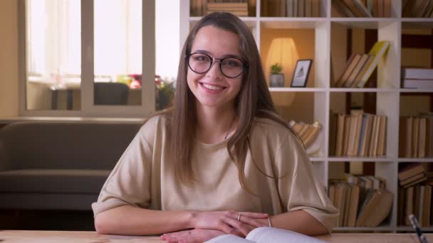 Close-up portret van jonge aantrekkelijke Kaukasische vrouwelijke student in glazen kijken naar camera glimlachend gelukkig binnenshuis in de Universiteitsbibliotheek — Stockvideo