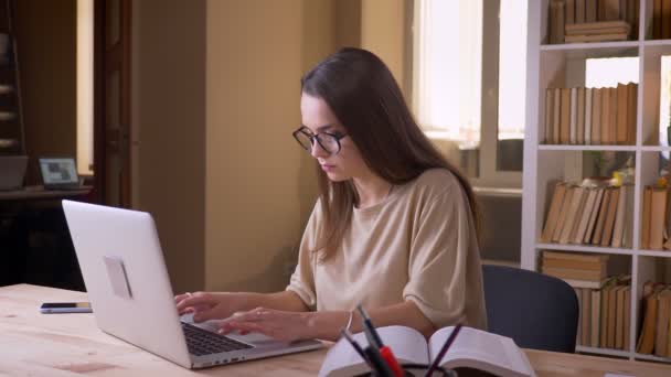 Portrait rapproché d'une jeune étudiante caucasienne attrayante utilisant l'ordinateur portable et lisant un livre à l'intérieur de la bibliothèque du collège — Video