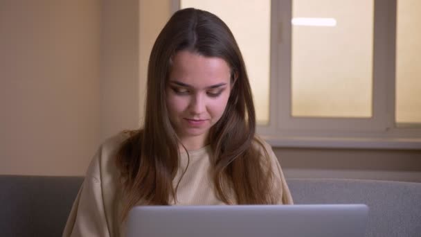 노트북을 사용하여 젊은 매력적인 백인 여성의 클로즈업 초상화와 행복하게 아파트의 소파에 앉아 웃음 — 비디오