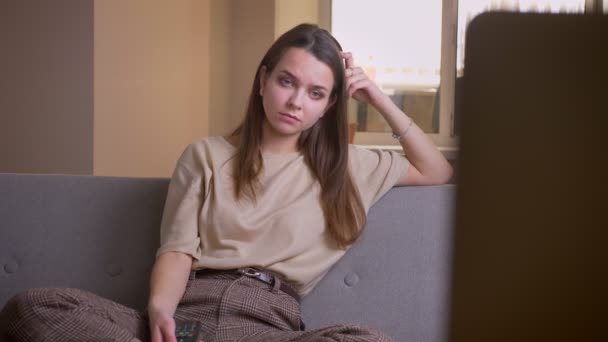 Primer plano retrato de joven atractiva mujer caucásica viendo la televisión sonriendo felizmente sentado en el sofá interior en el apartamento — Vídeos de Stock