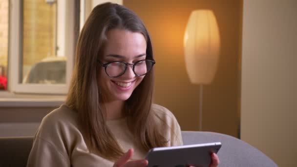 Крупним планом портрет молодої привабливої білої жінки в окулярах, використовуючи планшет, посміхаючись щасливо сидячи на дивані в квартирі — стокове відео