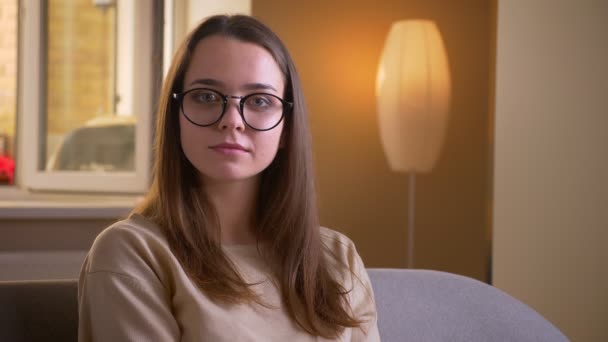 Retrato de close-up de jovem mulher branca atraente em óculos olhando para a câmera sentada no sofá dentro de casa em apartamento — Vídeo de Stock