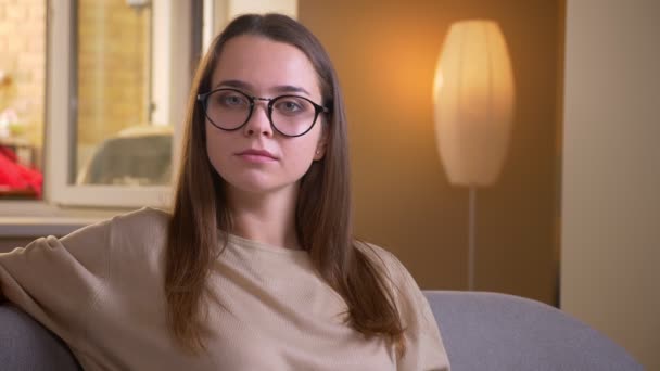 Closeup retrato de jovem mulher branca atraente em óculos sorrindo alegremente olhando para a câmera sentada no sofá dentro de casa em apartamento — Vídeo de Stock