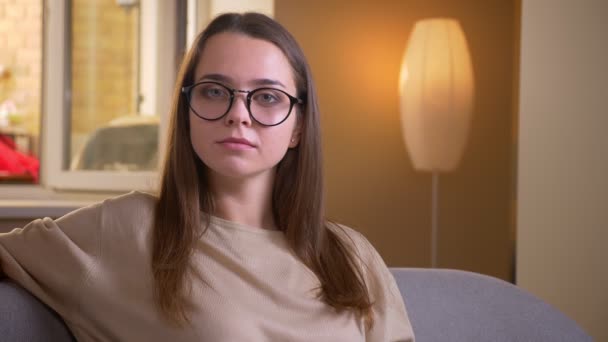 Primer plano retrato de joven atractiva hembra caucásica en gafas sonriendo felizmente mirando a la cámara sentada en el sofá interior en el apartamento — Vídeos de Stock