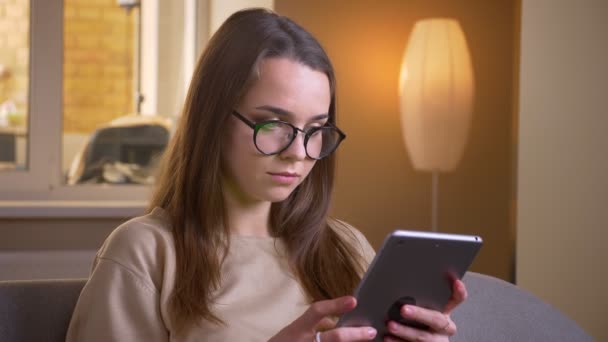 Крупный план портрета молодой привлекательной белой женщины в очках с помощью планшета и с зеленым экраном перед камерой, сидящей на диване в квартире — стоковое видео