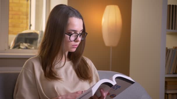 Primo piano ripresa di giovane studentessa attraente leggere un libro seduto sul divano al chiuso in un accogliente appartamento — Video Stock