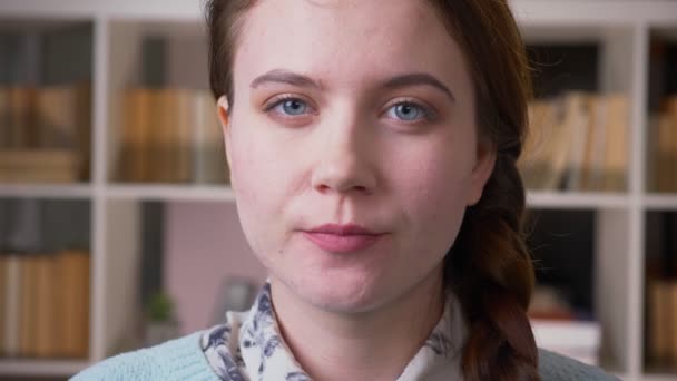 Nahaufnahme Porträt einer jungen hübschen Studentin, die in der Universitätsbibliothek drinnen in die Kamera blickt, mit Bücherregalen im Hintergrund — Stockvideo