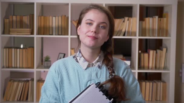 Retrato de cerca de una joven estudiante guapa sosteniendo un libro sonriendo mirando a la cámara de la biblioteca universitaria en el interior — Vídeos de Stock