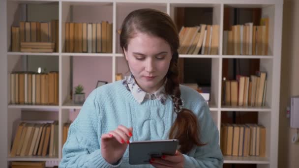 Κοντινό πλάνο του νεαρού όμορφη γυναίκα μαθητή μηνύματα στο δισκίο κοιτάζοντας την κάμερα στη βιβλιοθήκη του Πανεπιστημίου σε εσωτερικούς χώρους — Αρχείο Βίντεο