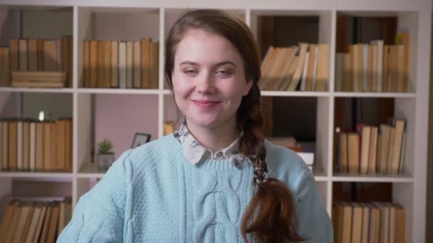 Retrato de primer plano de una joven estudiante bastante femenina que se muestra bien mirando la cámara en la biblioteca universitaria en el interior — Vídeos de Stock