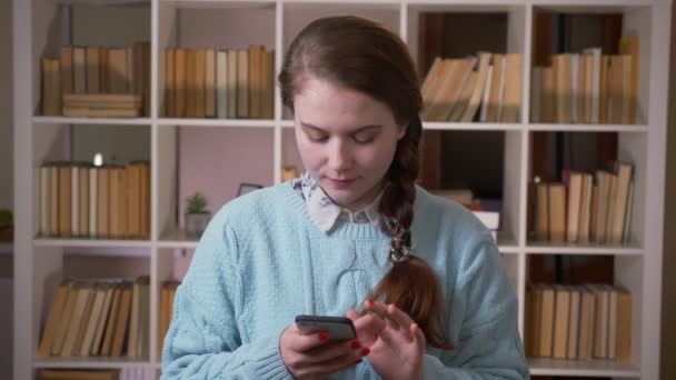 Портрет молодой красивой студентки с телефона, смотрящей на камеру в библиотеке университета в помещении — стоковое видео