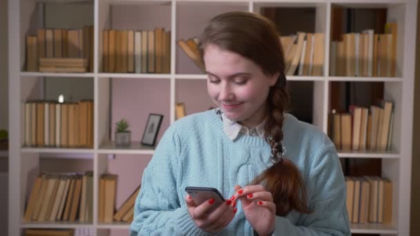 Крупный план портрета молодой красивой студентки, переписывающейся по телефону, смотрящей на камеру в библиотеке университета в помещении — стоковое видео
