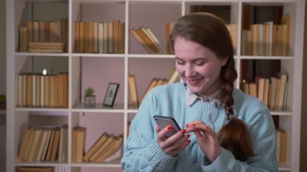 Närbild porträtt av unga vackra kvinnliga student surfar på telefonen tittar på kameran i Universitetsbiblioteket inomhus — Stockvideo