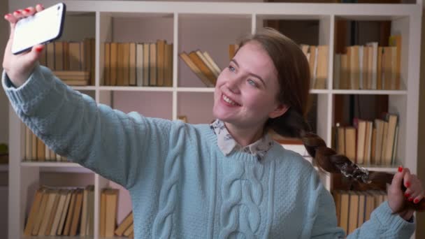 Gros plan portrait de jeune jolie étudiante prenant des selfies au téléphone en regardant la caméra dans la bibliothèque de l'université à l'intérieur — Video