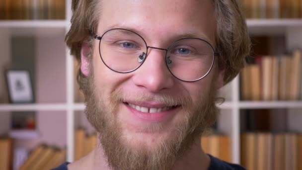 Lähikuva ampua aikuisten houkutteleva valkoihoinen mies opiskelija lasit hymyillen iloisesti katsoen kameran yliopiston kirjastossa sisätiloissa — kuvapankkivideo
