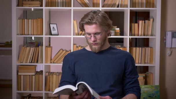 Nahaufnahme von erwachsenen attraktiven männlichen Studenten, die in der Universitätsbibliothek ein Buch lesen und in die Kamera schauen — Stockvideo