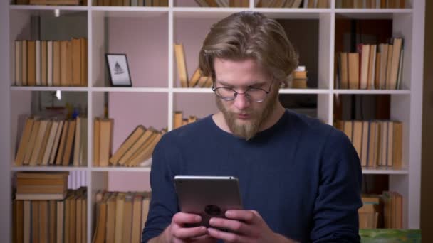 Sesión de primer plano de adulto atractivo estudiante masculino utilizando la tableta en la biblioteca de la universidad en el interior — Vídeos de Stock