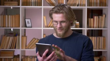 Tableti kullanarak ve üniversite kütüphanesinde kameraya yeşil renk ekranı gösteren yetişkin çekici erkek öğrencinin yakın çekim çekimi kapalı