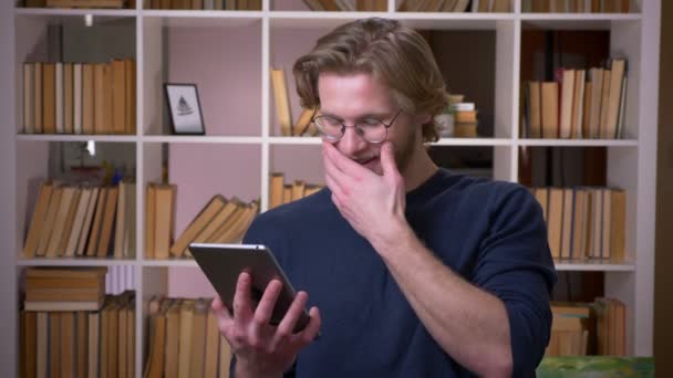 Съемки крупным планом взрослого привлекательного студента с помощью планшета, счастливо улыбающегося в университетской библиотеке в помещении — стоковое видео