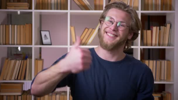 Крупним планом знімок дорослого привабливого студента-чоловіка, який показує великий палець і дивиться на камеру в університетській бібліотеці в приміщенні — стокове відео
