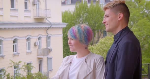 Tausendjähriges Paar steht auf Balkon und beobachtet grüne und sonnige Stadtansichten. — Stockvideo