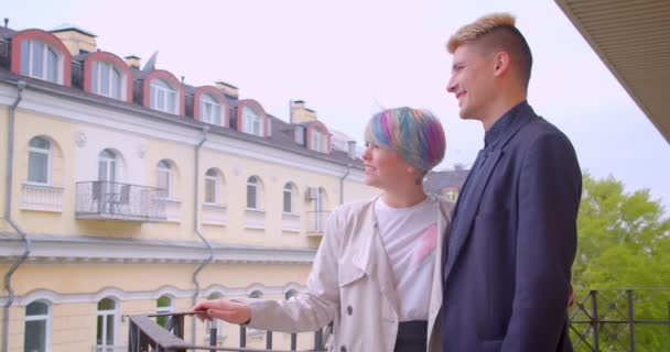 Millenial par står på balkongen tittar på grön och solig utsikt över staden vara glad och glad. — Stockvideo