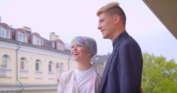 Millenial par står på balkongen tittar på grön och solig utsikt över staden och talar glatt vara lycklig. — Stockvideo