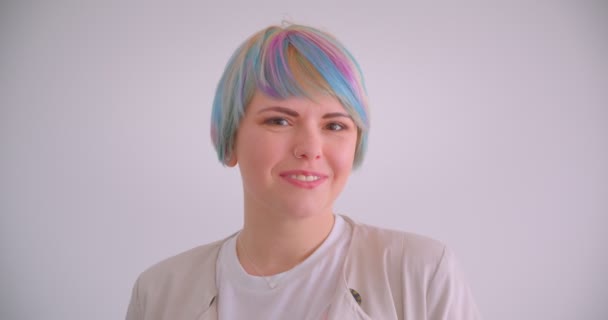 Garota caucasiana excêntrica com cabelos multicoloridos e sorrisos penetrantes timidamente na câmera no fundo branco . — Vídeo de Stock