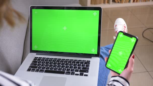 Крупный план вид сзади съемка женщины с помощью ноутбука и телефона с зеленым экраном сидит на диване в помещении в уютной квартире — стоковое видео