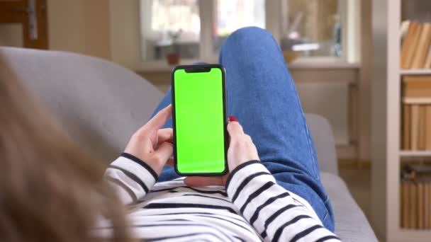 Крупним планом задній перегляд знімання жінки за допомогою телефону з зеленим екраном, що лежить на дивані в приміщенні в затишній квартирі — стокове відео
